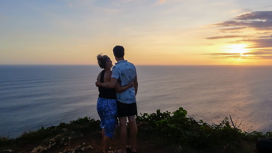 Couple standing at the cliffs edge hugging. The sky is orange.